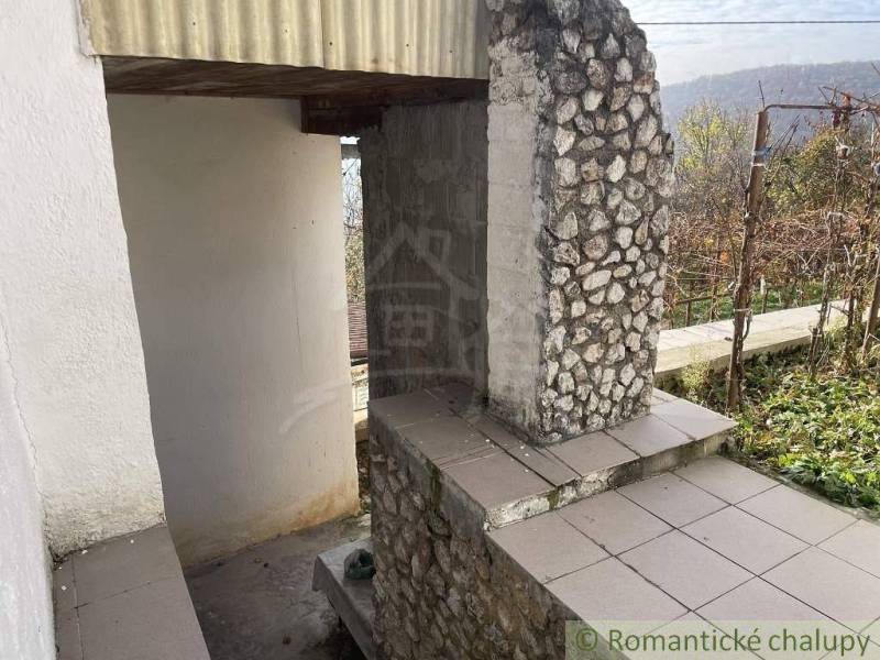 A stone and brick wall in front of a cottage surrounded by vineyards and a garden.