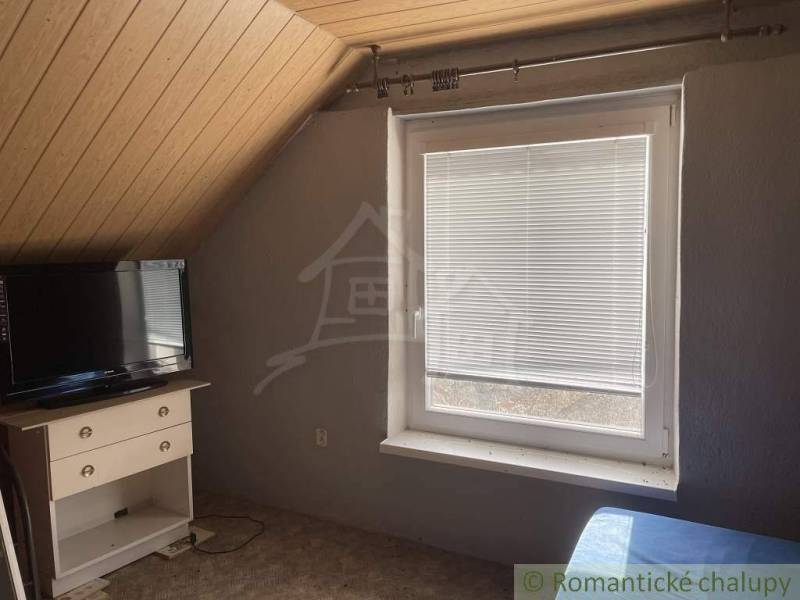 A television and a dresser in the attic room of a cottage with a window and a wooden ceiling.
