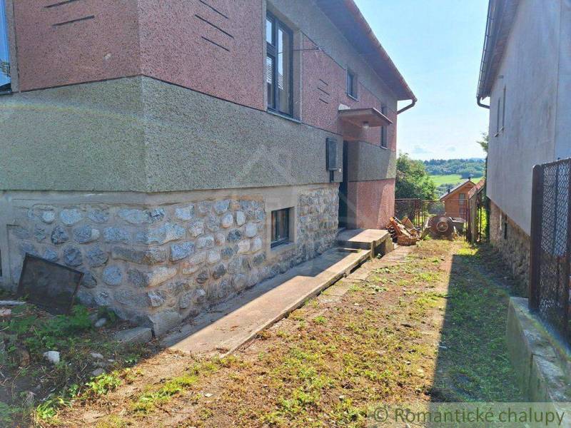 A family house with a stone foundation, next to a fence and a concrete road in a rural setting.