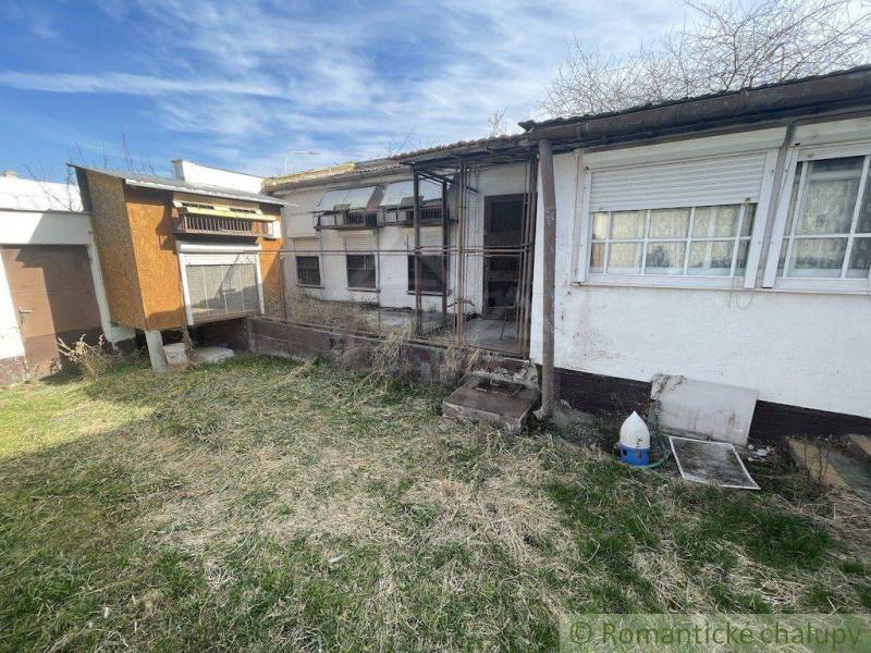 A yard with a neglected family house and an annex, surrounded by a lawn and a clear sky.
