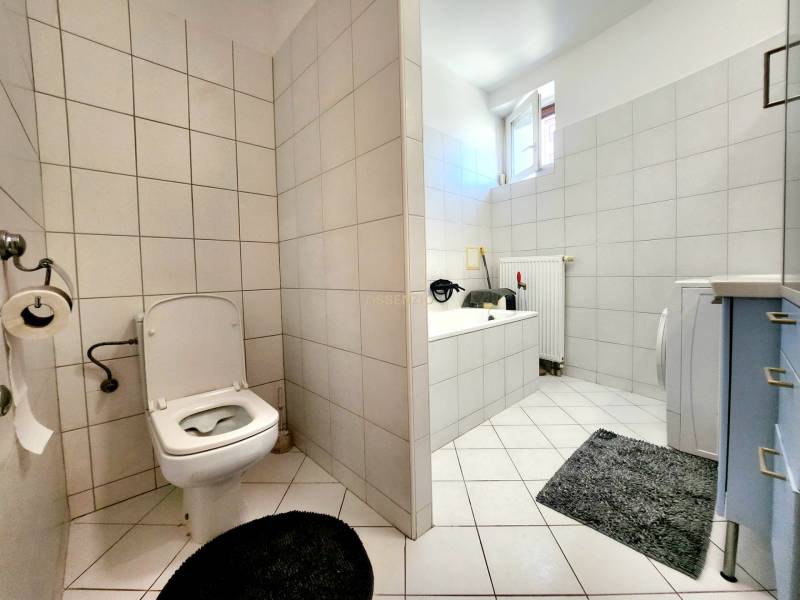 A bathroom in a studio apartment with a bathtub, washing machine, and toilet, tiled with light-colored tiles.
