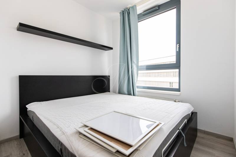 Bedroom with a bed and a shelf in a 2-room apartment, light curtains and a large window.