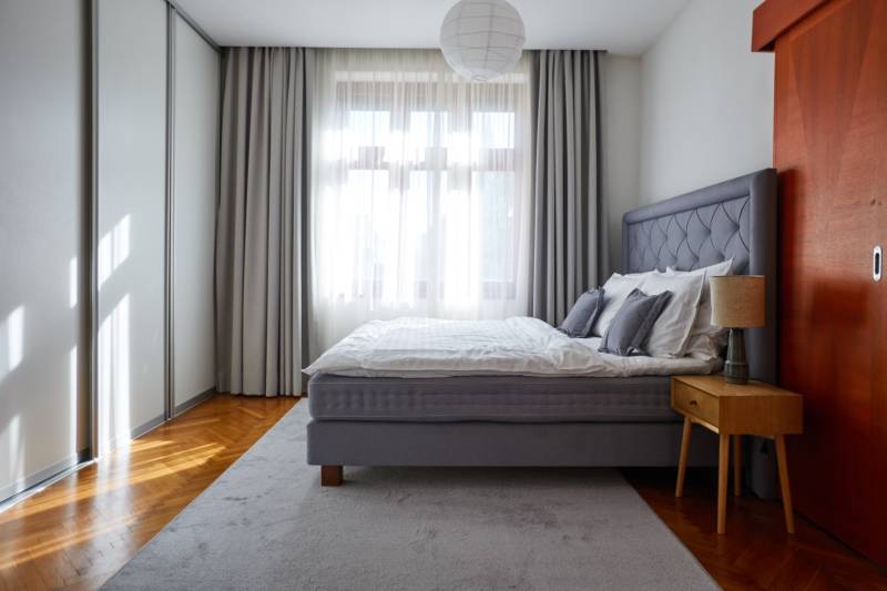 A bright bedroom with a gray bed, a nightstand, and large windows in a 4-room apartment.