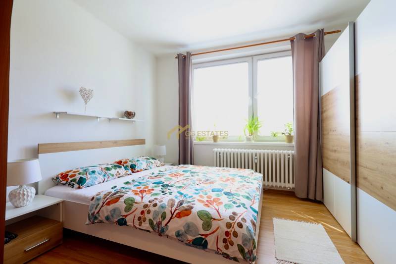 A bedroom in a two-room apartment with a large window, curtains, and flowers on the windowsill.