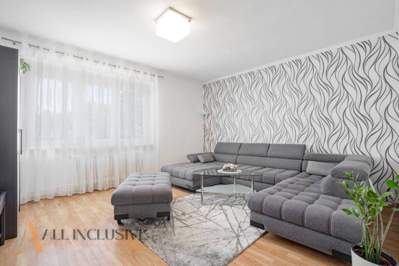Living room of a 2-room apartment with a comfortable gray sofa, patterned wallpaper, and wooden flooring.