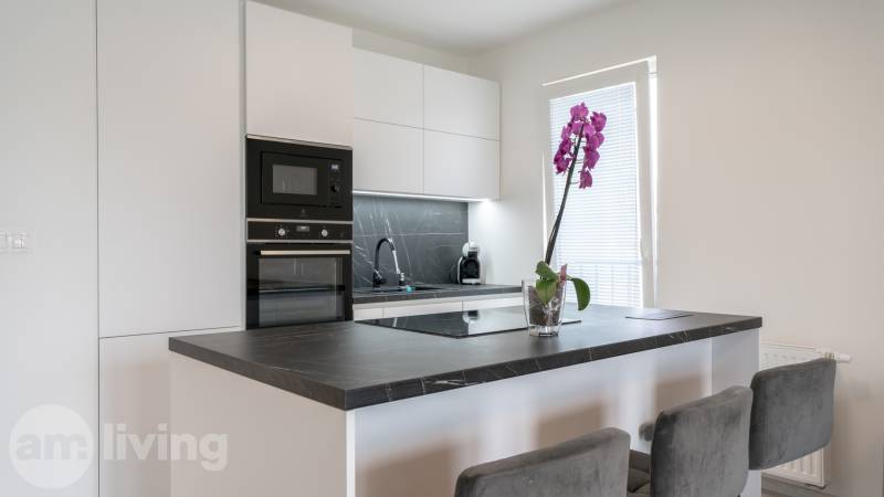 A kitchen in a 3-room apartment with built-in appliances, a black countertop, and a pink orchid.