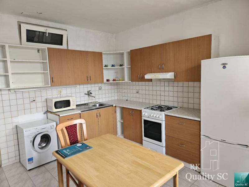 Kitchen area of a 2-room apartment with a refrigerator, washing machine, and stove, wooden table with a chair.