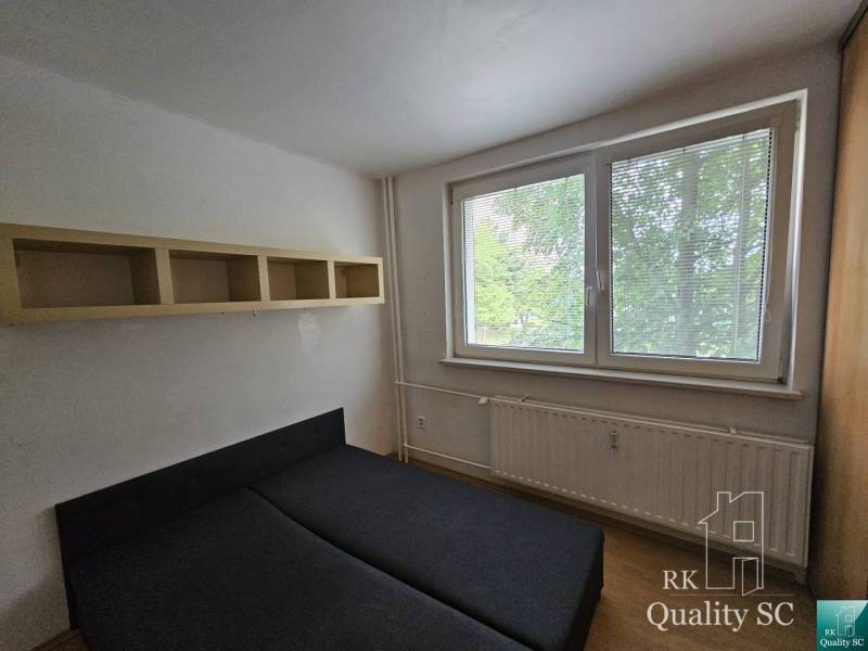 A bedroom in a 2-room apartment with a bed, a shelf, and a window with a view of greenery.