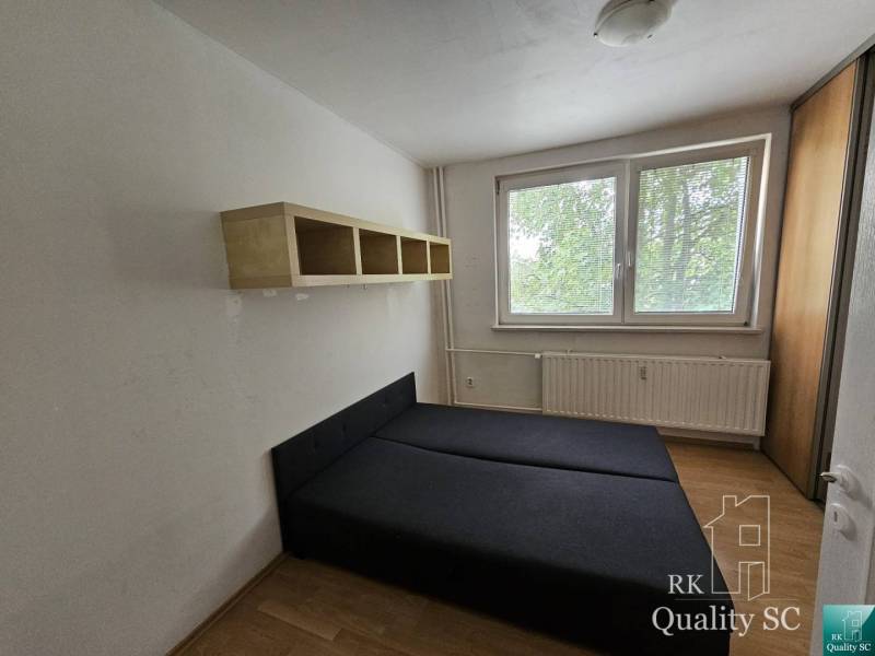 A bedroom with a bed and a shelf in a two-room apartment. Windows with a view of greenery.