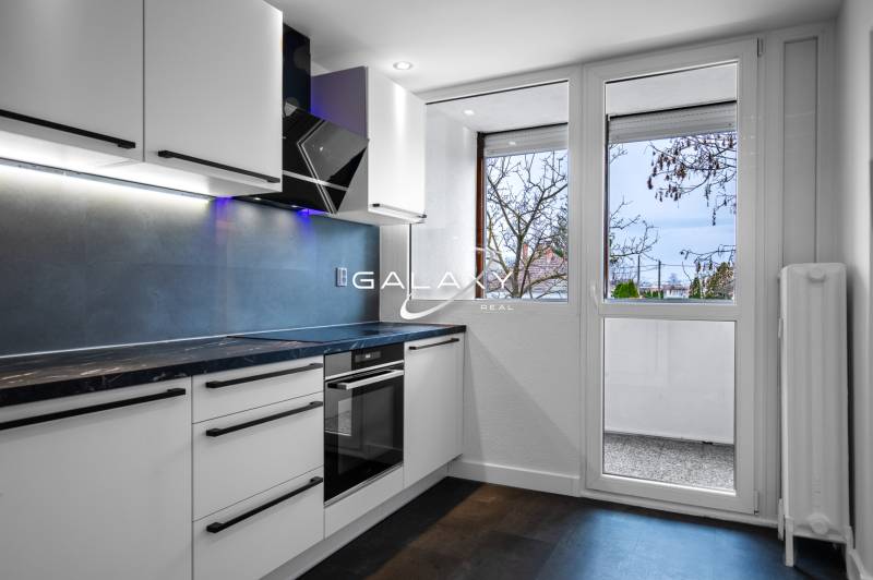 A kitchen in a 3-room apartment with white cabinets, a black cooking surface, and a window.