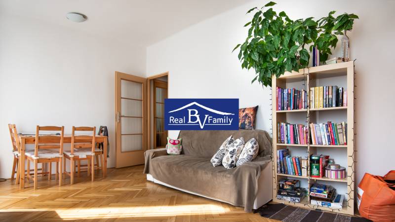 Studio apartment with wooden flooring, a sofa, a bookshelf, and a dining table.