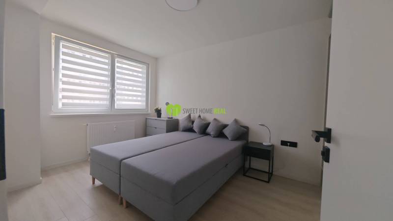 Bedroom in a studio apartment with a double bed, nightstands, and a window with blinds.