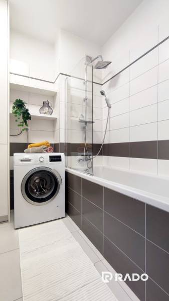 A bathroom in a 2-room apartment with a washing machine, bathtub, shelves, and a decorative plant.