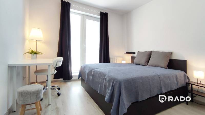 Bright bedroom in a 2-room apartment with a bed, table, and window with curtains.