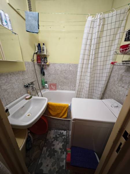 A bathroom in a 2-room apartment with a washing machine, bathtub, sink, and light tiles.