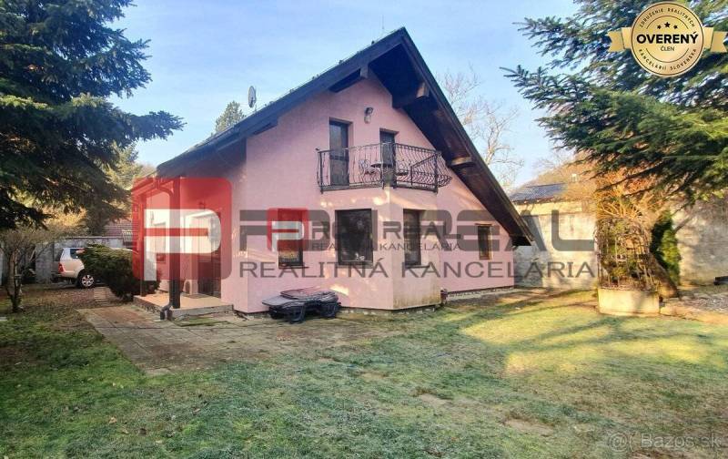 A family house in Píla with an attic roof, a balcony, and a garden surrounded by trees.