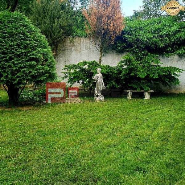 The garden of a family house in the village of Píla with a lawn, a statue, and a bench by the wall.
