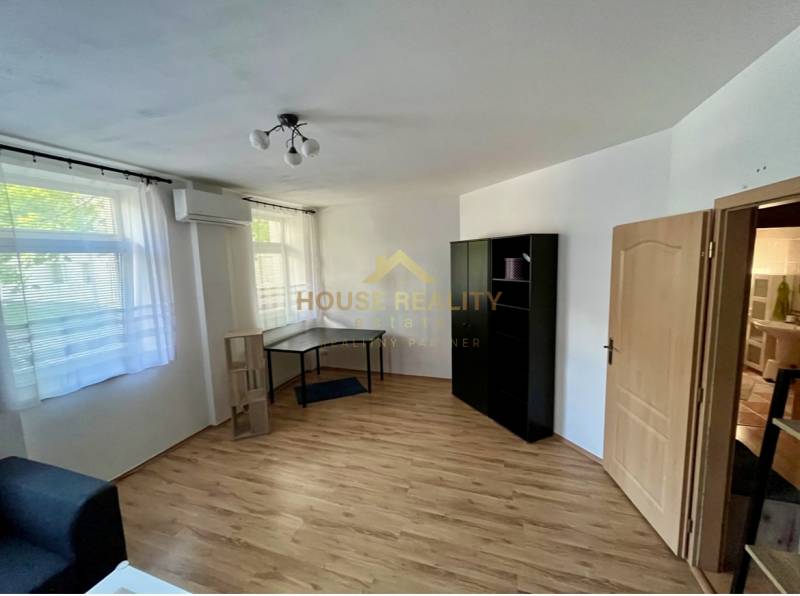 A room in a 2-room apartment with wooden flooring, air conditioning, furniture, and windows with blinds.