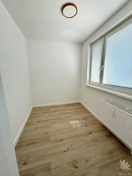 A room with light walls, wooden flooring, and a large window in a 2-room apartment.