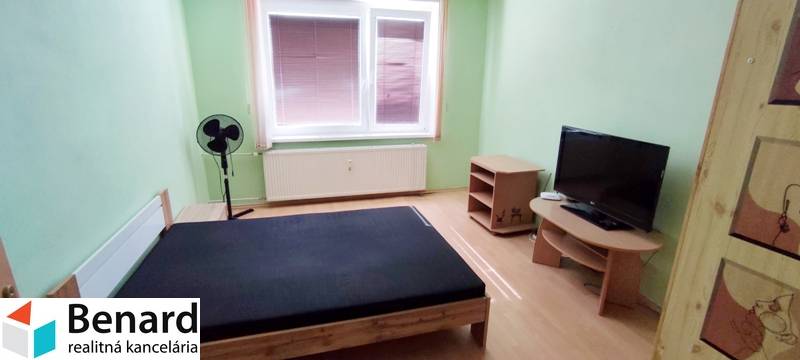 Bedroom in a two-room apartment with a bed, television, and a fan in the corner of the room.