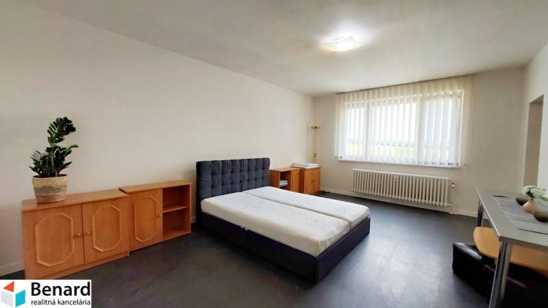 A simply furnished room with a bed, a dresser, and a large window in a studio apartment.