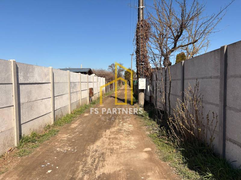 A narrow path leads between concrete fences, accompanied by trees and a blue sky.