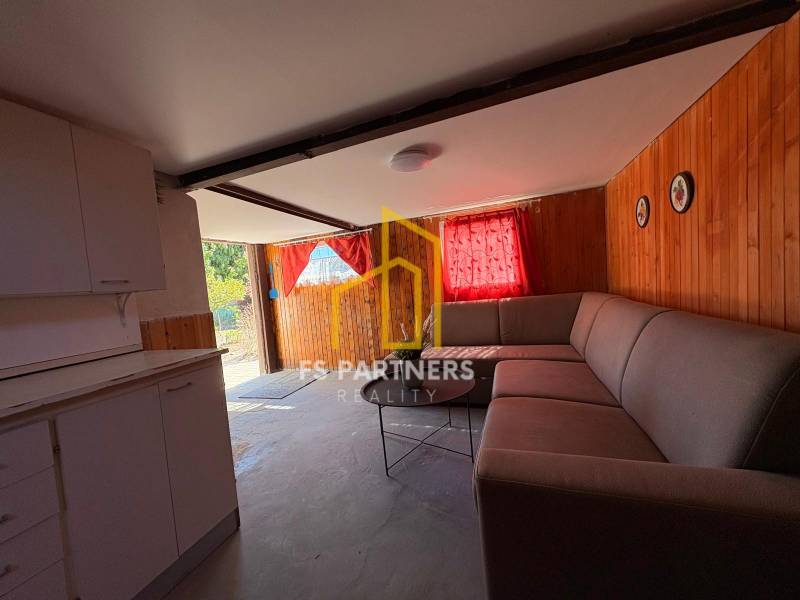 An interior with a corner sofa and a kitchen unit, wooden paneled walls, red curtains.