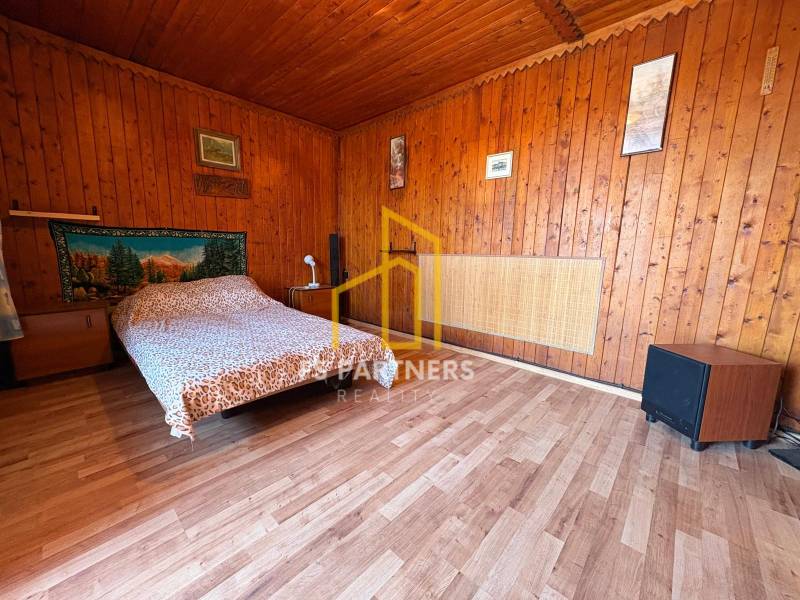 Wooden interior with a bed, decorative fabric on the wall, paintings, and simple furniture.