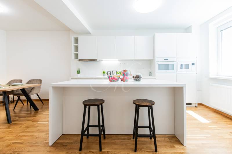 A kitchen in a 3-room apartment with a white kitchen unit, bar stools, and a wooden floor.