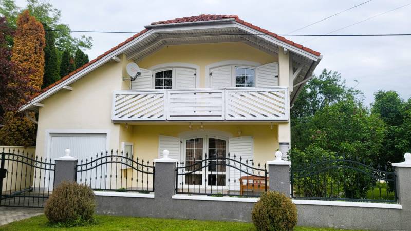 A family house in Dunasziget with white shutters and a balcony, surrounded by greenery.