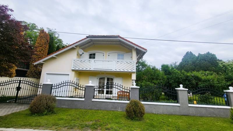 Yellow family house in Dunaszige, with a balcony and a garden, enclosed by a black fence.