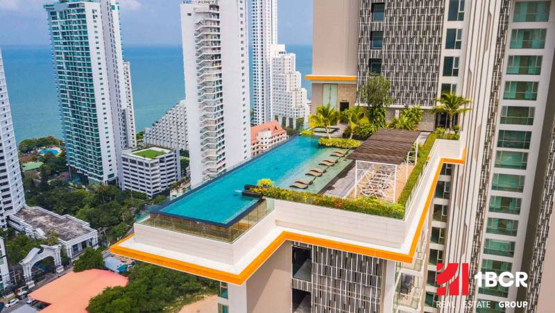 Apartment construction in Pattaya with a rooftop pool, with the sea and high-rise buildings in the background.