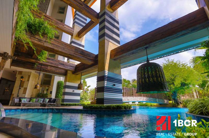 A luxurious pool with wooden beams, surrounded by greenery, falls under the construction of apartments.