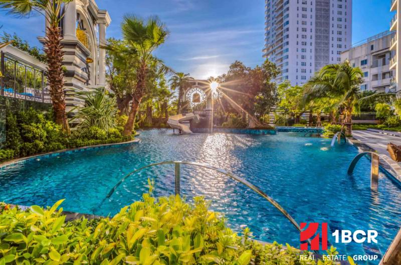 Apartment construction in Pattaya with a pool surrounded by greenery and palm trees in a sunny environment.