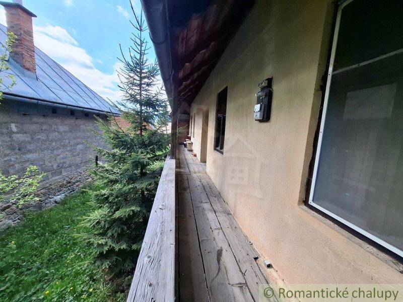A wooden walkway next to a family house in Pohorelá, surrounded by greenery and a neighboring building.