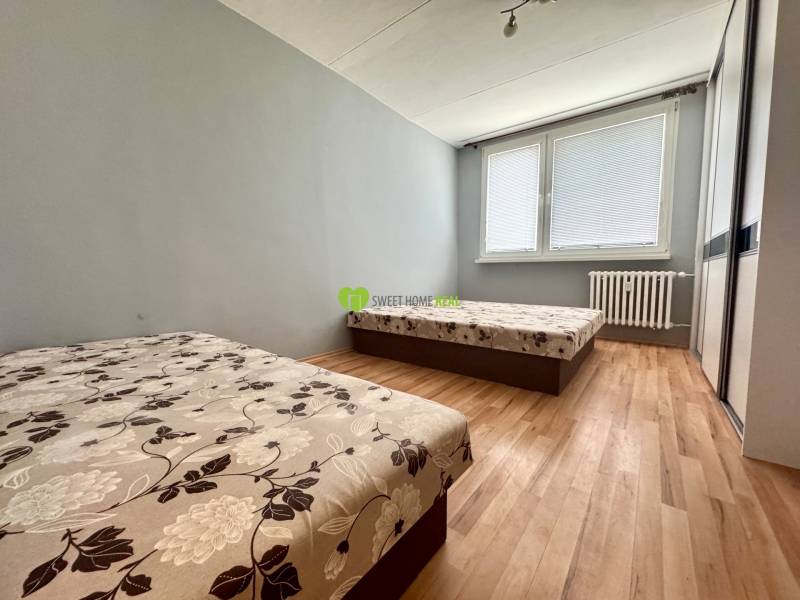 Two beds, wooden floor, and a window with blinds in a three-room apartment.