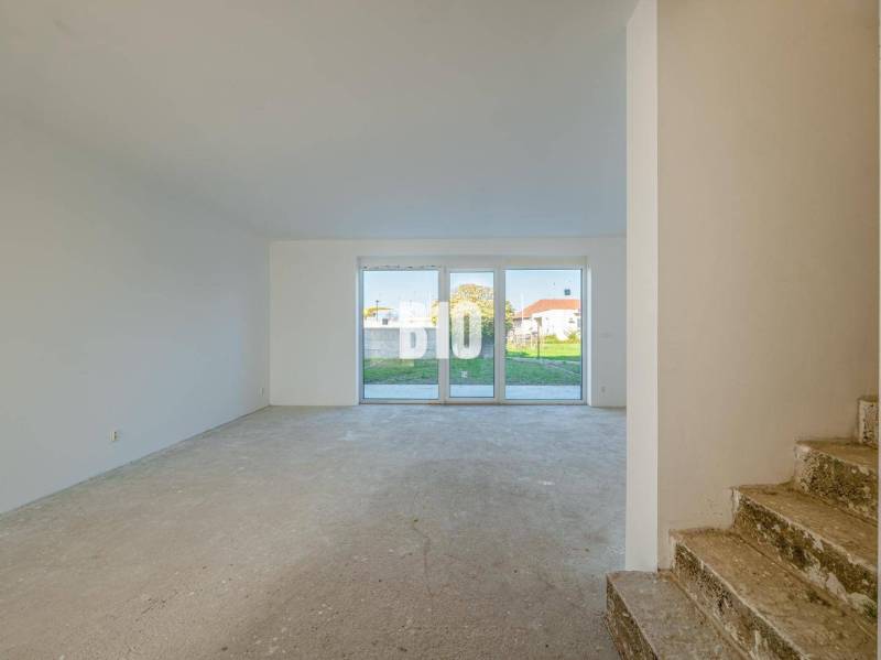 A space in a family house with large windows and a concrete floor, stairs in the background.
