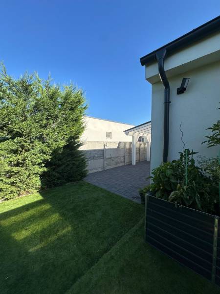 A lawn and a paved walkway at a family house in Gabčíkovo with shrubs and a clear sky.