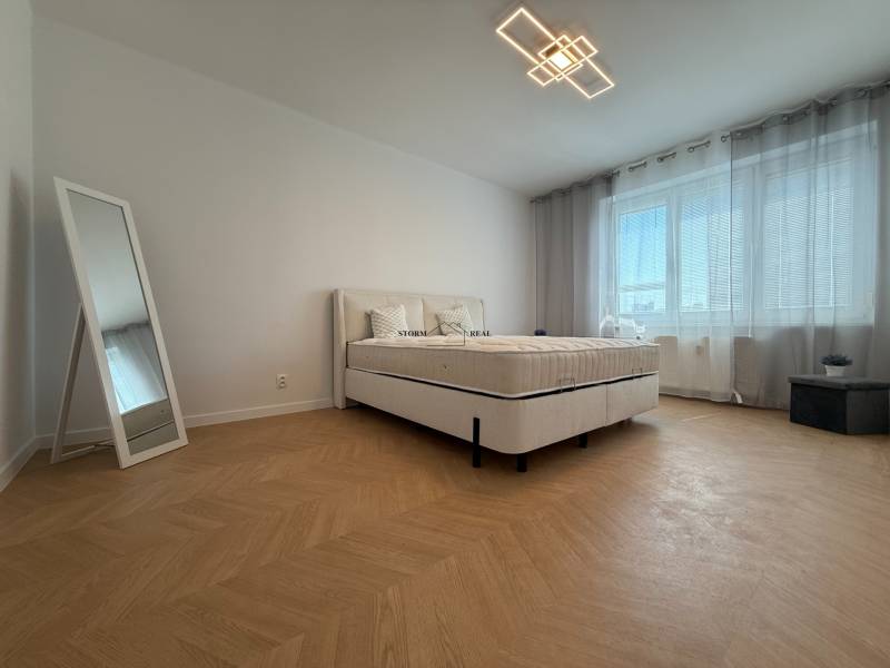 A bedroom in a 3-room apartment with a bed, a mirror, and a large window with blinds.