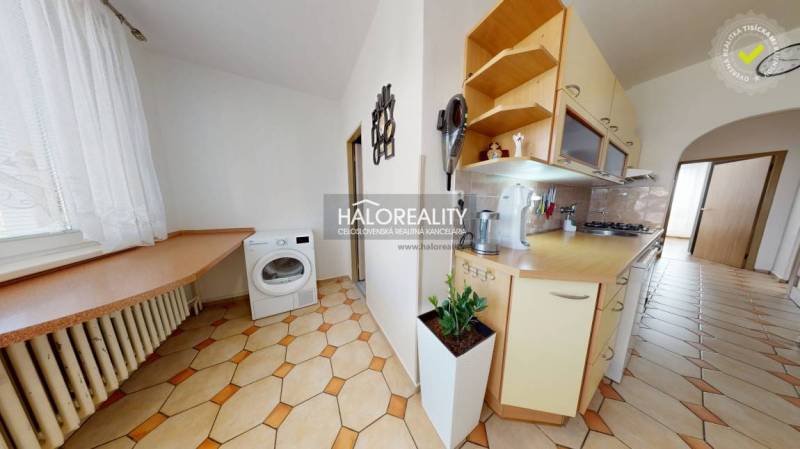 A kitchen in a 3-room apartment with a washing machine, kitchen unit, and a plant on the floor.