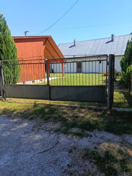 A gate and a wooden cottage in Tužina, surrounded by a lawn and coniferous trees.