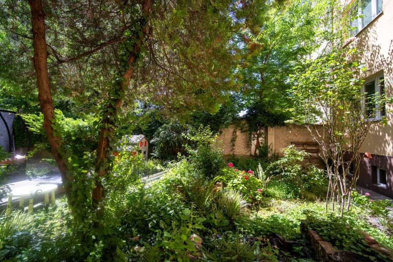 A garden full of greenery with a wooden house by a 3-room apartment, surrounded by trees.