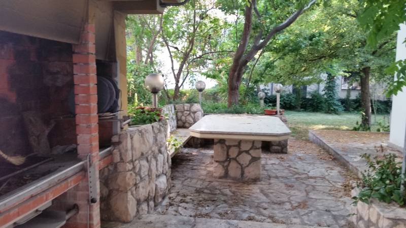 The courtyard of a family house in Vrsi with a stone table and a fireplace surrounded by greenery.