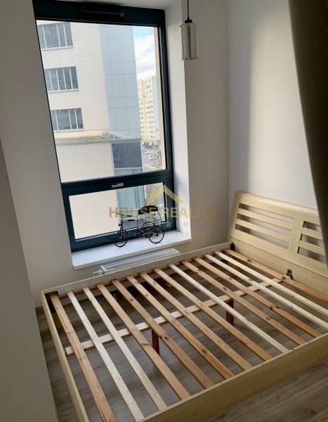 Bright bedroom in a 2-room apartment with a large window and an empty wooden bed frame.