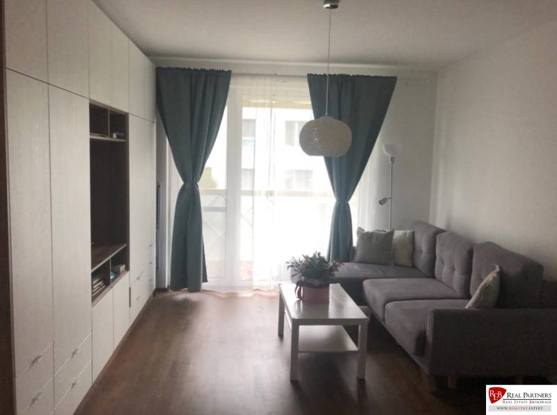 Living room in a two-room apartment with a sofa, furniture, curtains, and a table with a flower.