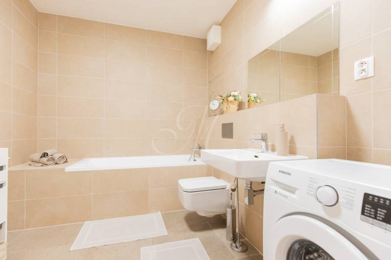 A bathroom with light tiles, a bathtub, a sink, and a washing machine in a studio apartment.