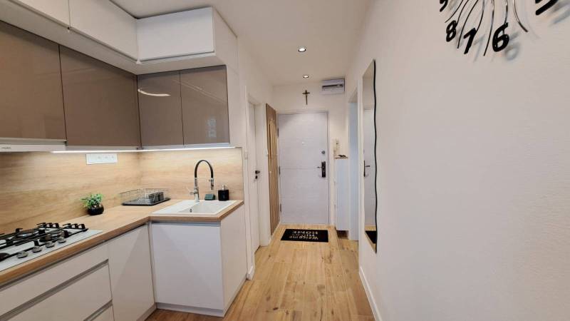 A kitchen in a 2-room apartment with white cabinets, a wooden countertop, and a large clock.