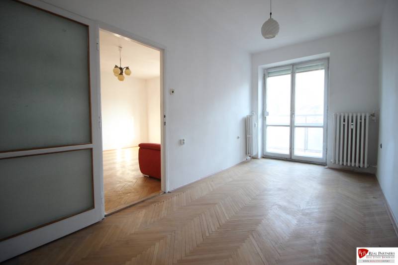 An empty room with parquet flooring leading to a room with a red couch in a 3-room apartment.