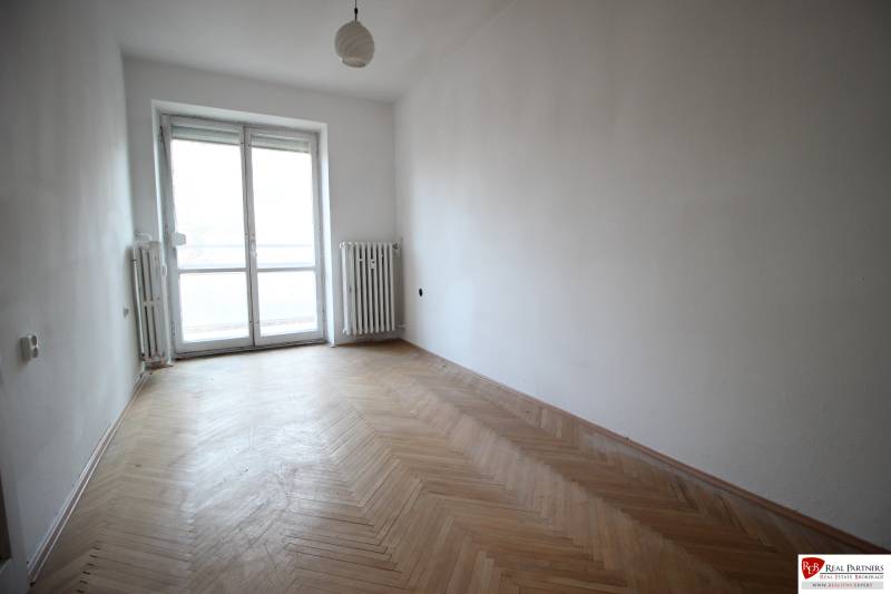 An empty room with parquet flooring in a 3-room apartment, with a large window and radiators.