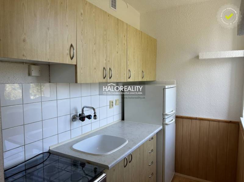 A kitchen in a studio apartment with wooden cabinets and a refrigerator, white tiles.